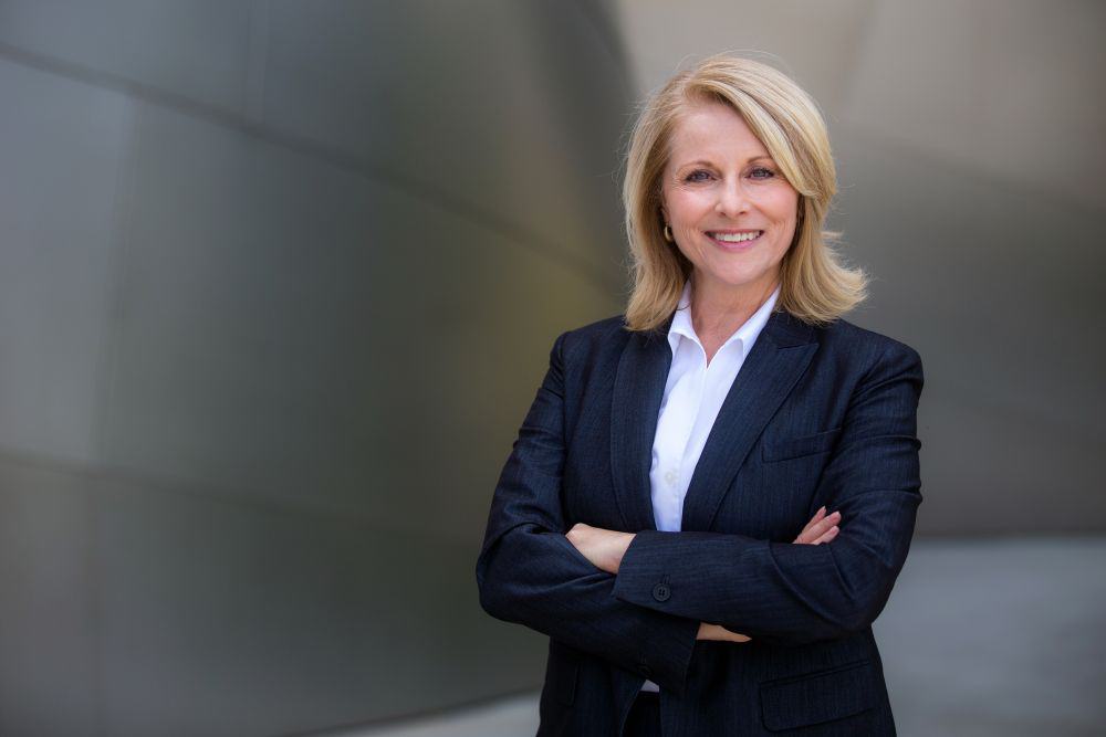 A woman stands with her arms crossed, wearing a dark suit and white blouse
